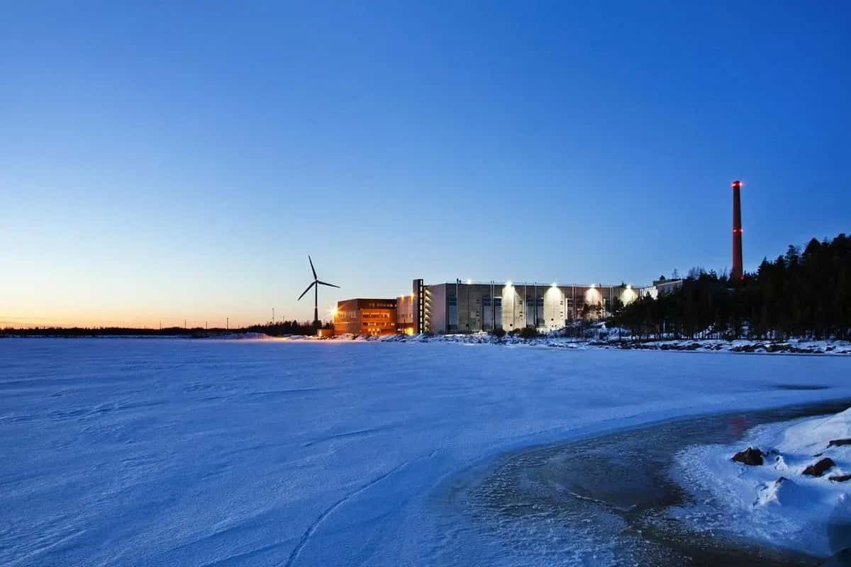 Vinterlandskap med snö, byggnader, vindkraftverk och röd skorsten. Blå himmel