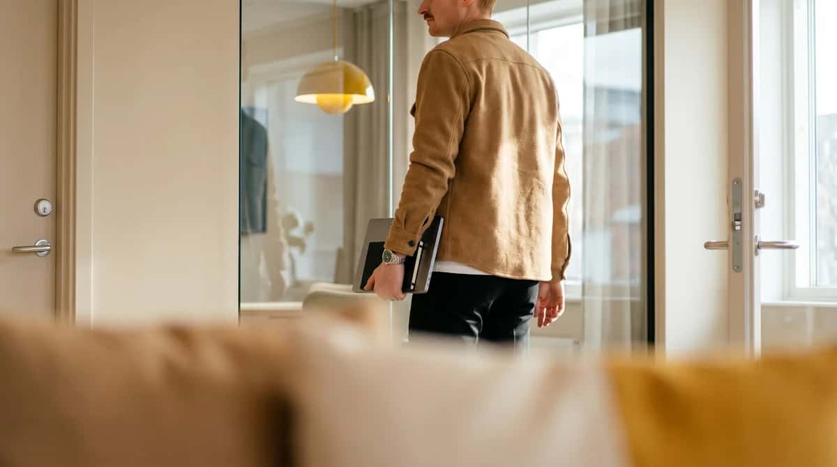 En man i beige jacka sedd bakifrån bär en bärbar dator och en anteckningsbok