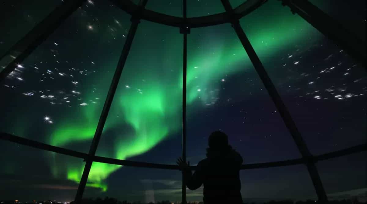 Person ser grönt norrsken och stjärnor genom ett kupolformat fönster