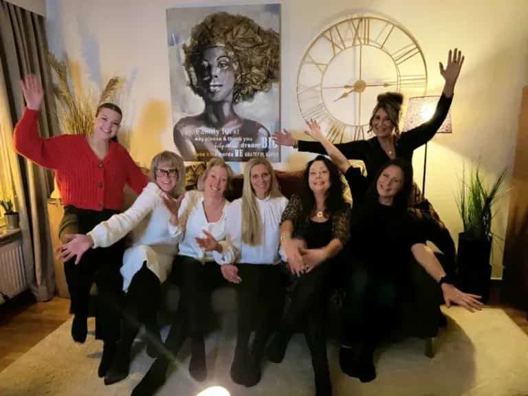 Sju leende kvinnor sitter i en soffa och vinkar mot kameran. En tavla och en klocka syns i bakgrunden