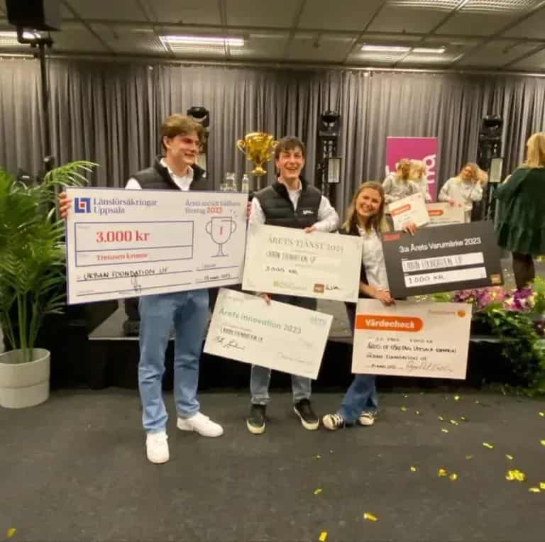 Sex personer poserar med blommor och stora checkar med "Collect UF". Logotyper i bakgrunden